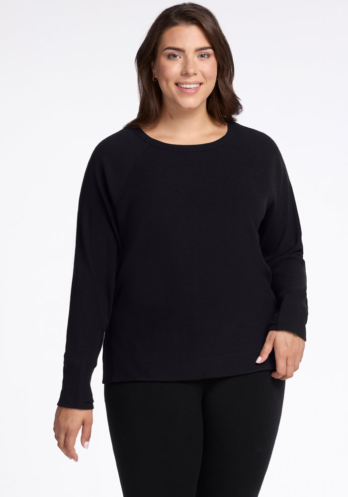 A woman with long brown hair smiles, wearing the Woolx Ainsley Sweater in black, crafted from temperature-regulating Merino wool, along with matching pants, as she stands against a plain white background. | Juliana is 5'10", wearing a size XL