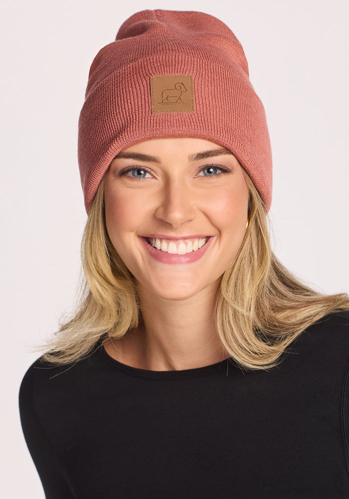 A smiling woman with blonde hair wears the Woolx Baylor Beanie in Brick Dust, featuring a small square patch, and a black long-sleeve shirt, standing against a plain light background.
