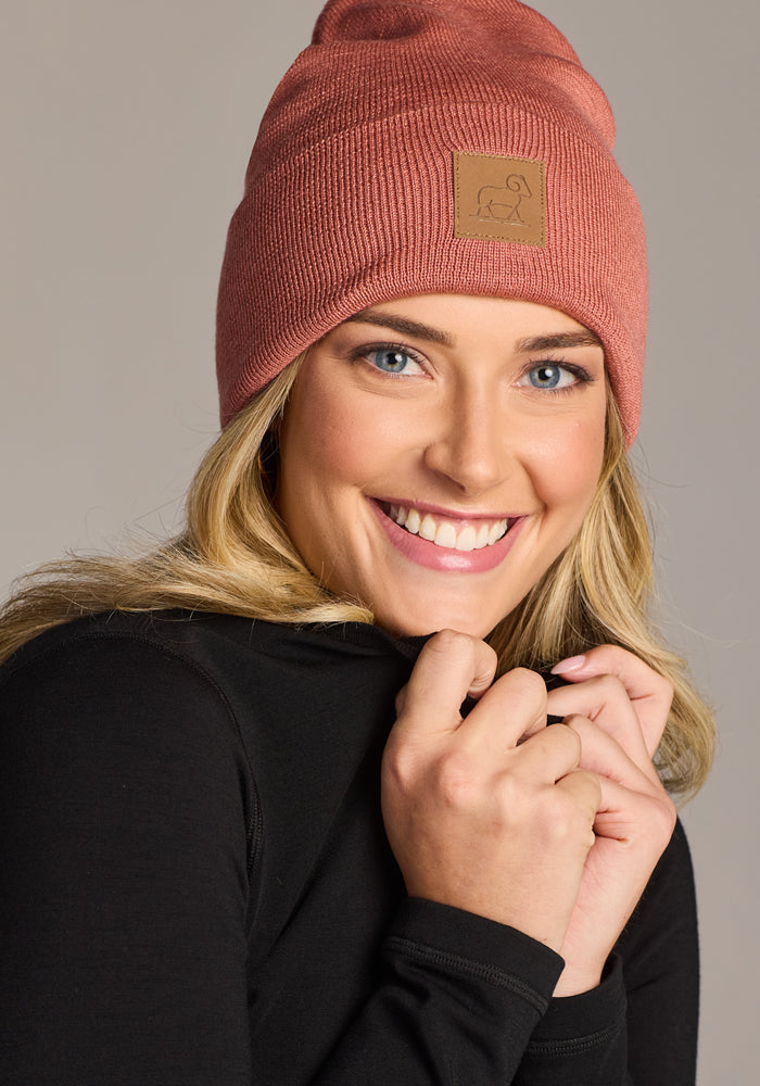 A smiling person wears the Woolx Baylor Beanie in Brick Dust and a black long-sleeve shirt, posing with hands near their face against a neutral background.