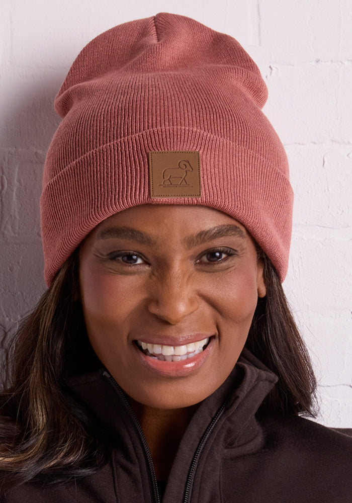 A woman smiles in front of a white brick wall, ready for chilly days in her warm Woolx Baylor Beanie - Brick Dust, made of merino wool with a small square patch featuring a simple ram outline, paired with a dark zip-up jacket.