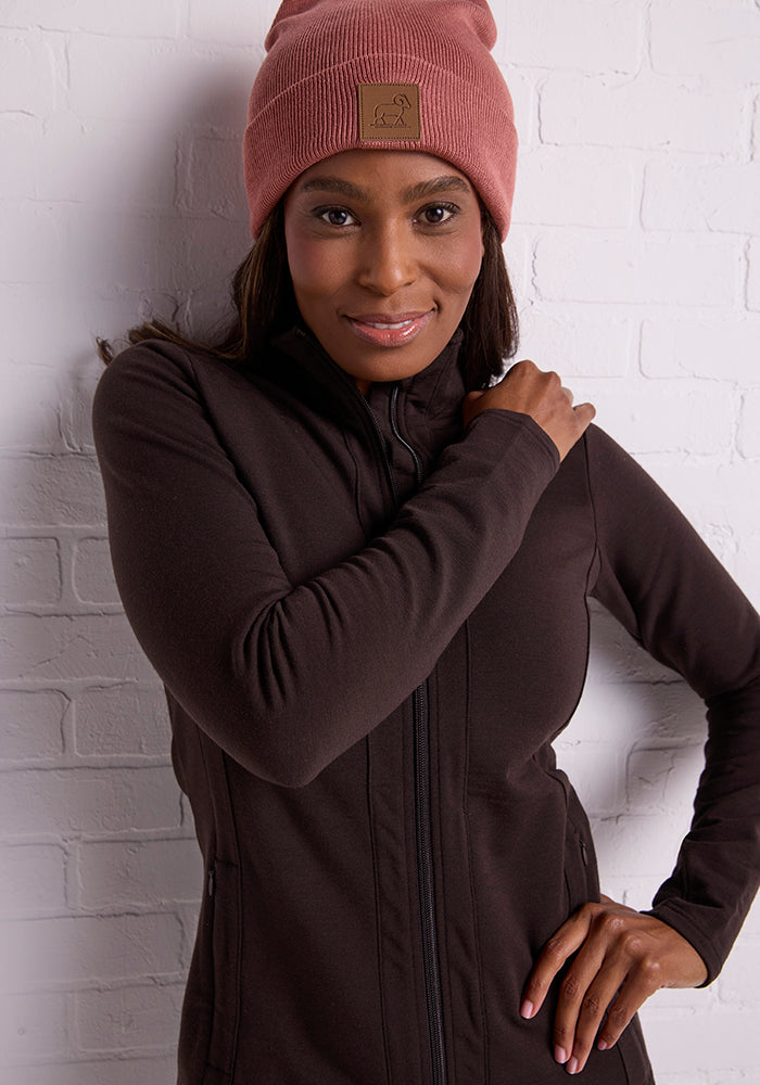 A woman wearing the Woolx Baylor Beanie in Brick Dust and a dark zip-up jacket stands against a white brick wall, smiling softly with one hand resting on her shoulder.