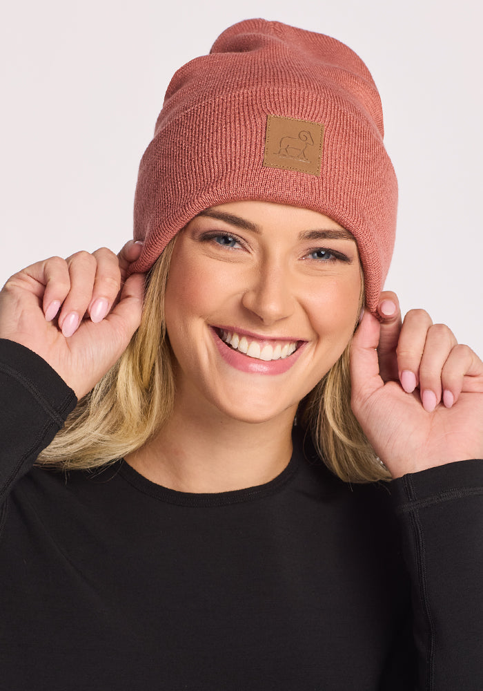 A smiling blonde woman wears the Woolx Baylor Beanie in Brick Dust and a black long-sleeve shirt, holding both sides of her warm winter hat against a light background.