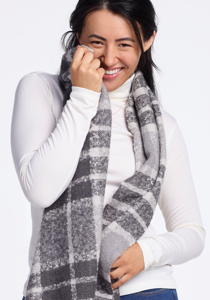 A person in a white turtleneck smiles warmly while holding the Bradie Scarf in Dark Grey Plaid by Woolx up to their cheek against a plain background.