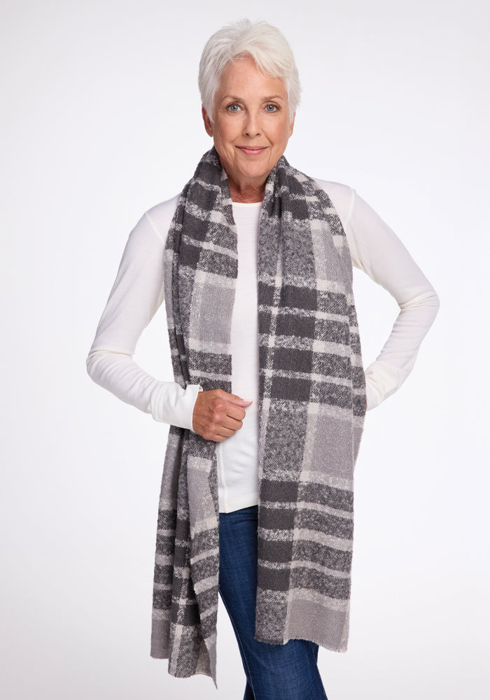 An older woman with short gray hair smiles warmly while wearing the Woolx Bradie Scarf in dark grey plaid, paired with a white long-sleeve top and blue jeans. She exudes confidence as she stands against a simple white background.