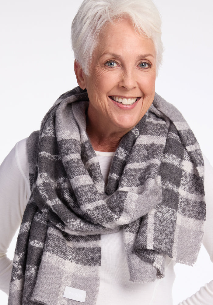 A smiling person with short white hair is wearing the Woolx Bradie Scarf in Dark Grey Plaid over a white top, posing against a plain background.