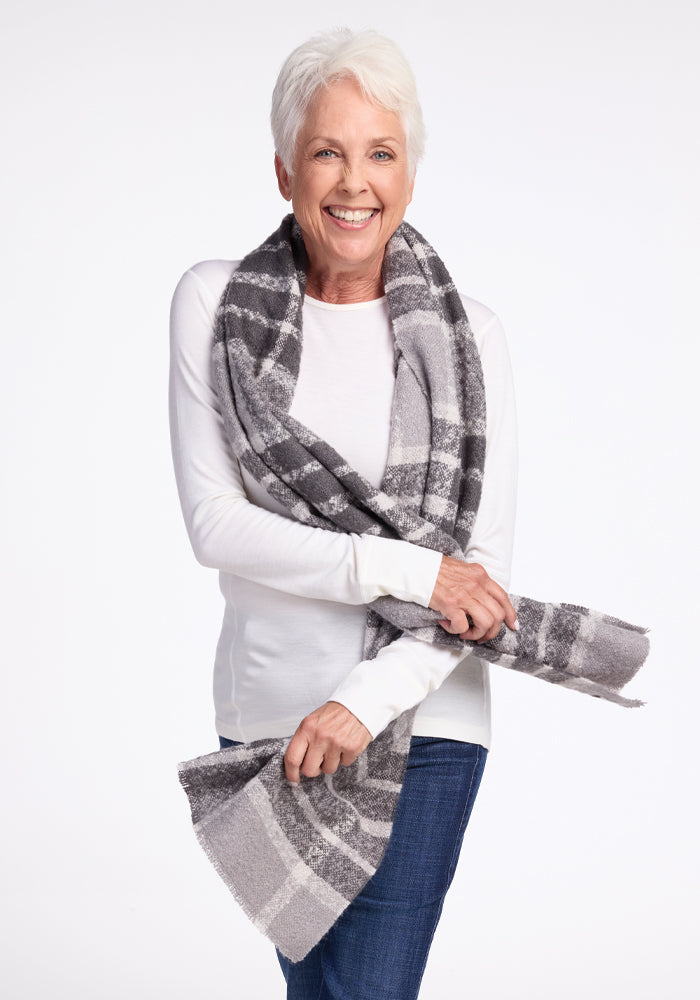 A smiling person with short white hair is wearing a white long-sleeve shirt and blue jeans. They have the Bradie Scarf in Dark Grey Plaid from Woolx wrapped around their neck, holding the ends with their hands. The background is plain white.