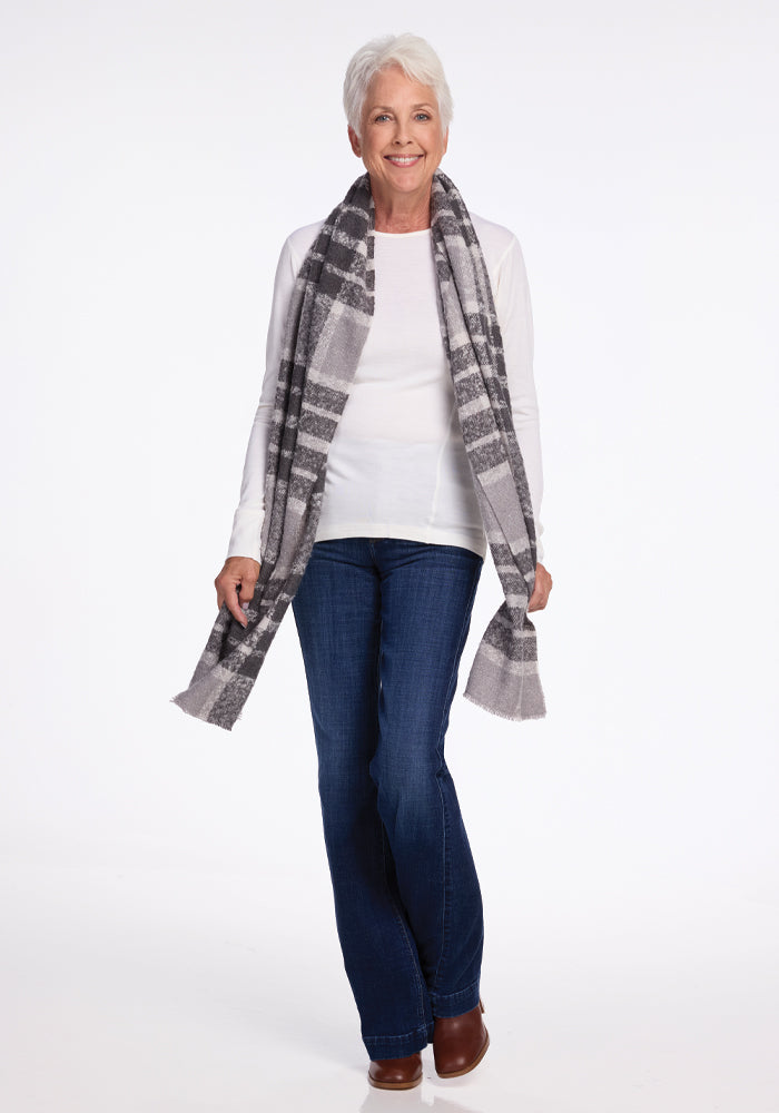A person with short white hair is smiling, wearing a white long-sleeve shirt, blue jeans, and brown shoes. The Woolx Bradie Scarf in dark grey plaid is draped around their neck. The background is plain white.