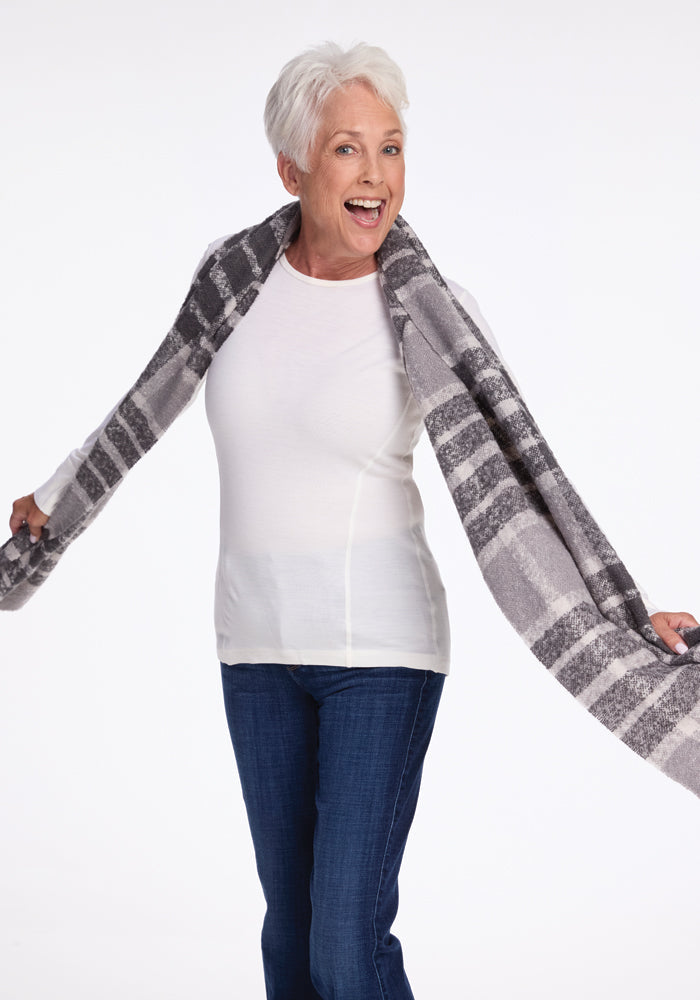 An older adult with short gray hair, dressed in a white long-sleeve shirt and blue jeans, smiles broadly while holding the Bradie Scarf in Dark Grey Plaid by Woolx extended behind her against a plain white background.