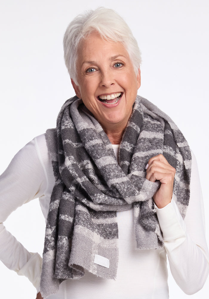 A smiling older woman with short white hair wears a cozy, dark grey plaid Bradie Scarf from Woolx over a white long-sleeve top. She stands against a plain white background, exuding warmth and happiness.