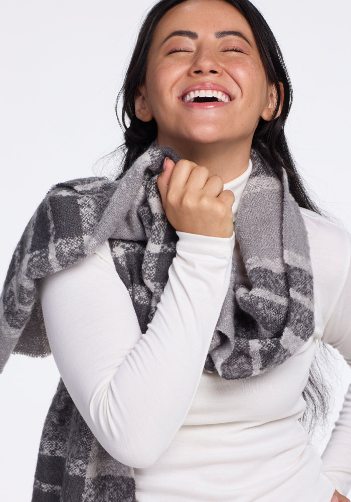A person with long dark hair is laughing joyfully while wearing a white long-sleeve shirt and the Woolx Bradie Scarf in Dark Grey Plaid. The background is plain white.