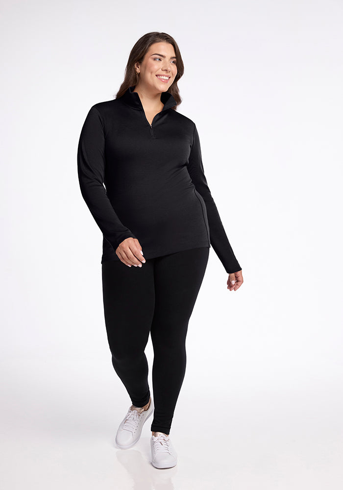 A person in the Woolx Brooke 1/4 Zip OLD, a moisture-wicking long-sleeved black top, paired with black leggings walks confidently against a plain white background. They have light brown hair and are smiling, with their left hand slightly raised. Wearing white sneakers, they exude casual style and comfort.