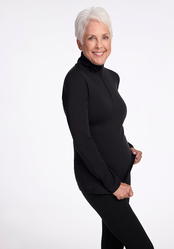 An older woman with short white hair is smiling at the camera, dressed in the athletic fit Brooke 1/4 Zip OLD top and matching black pants from Woolx, crafted from moisture-wicking fabric. The background is plain white.