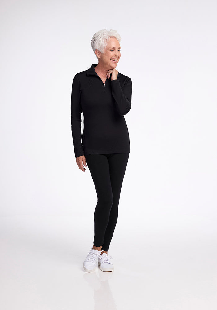 An older adult with short white hair is depicted wearing Woolx's Brooke 1/4 Zip OLD, a moisture-wicking black long-sleeve top, along with black leggings. They are smiling while looking to the side, styled in white sneakers and set against a plain white background.