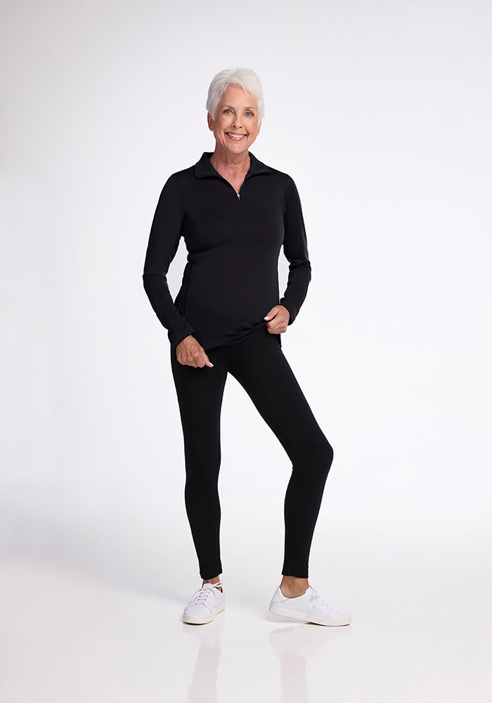 A smiling person with short white hair stands against a white background, wearing Woolx's Brooke 1/4 Zip OLD athletic-fit long-sleeved black top and black pants. They are holding the hem of their top and wearing white sneakers.