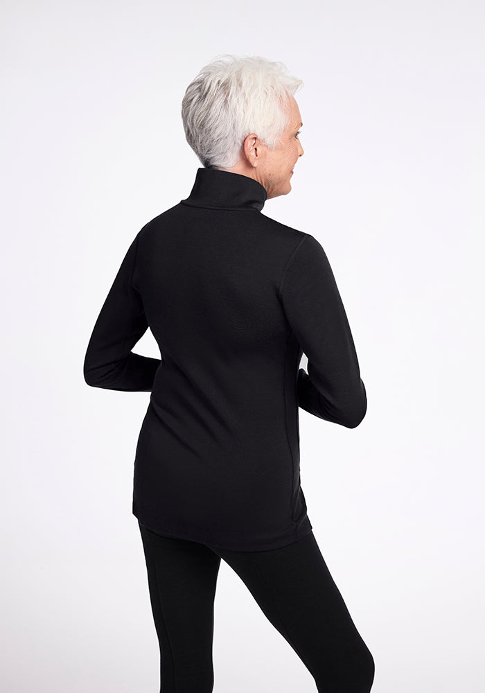 An older adult with short white hair is seen wearing the Brooke 1/4 Zip OLD from Woolx in black and matching pants. They are standing and facing away, with their hands resting on their hips against a plain white background.