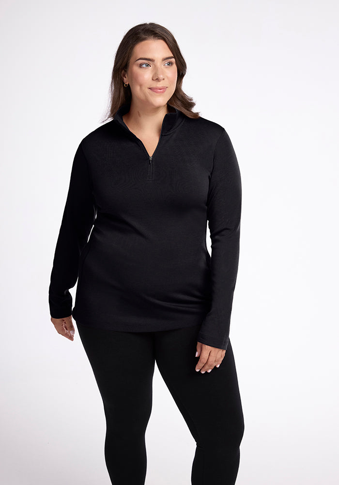 A person dressed in the Woolx Brooke 1/4 Zip OLD, a black top and pants, stands against a plain white background. The elegant athletic fit features moisture-wicking fabric and a zippered collar. They smile softly while looking to the side, showcasing the stylish design. | Juliana is 5'10", wearing a size XL