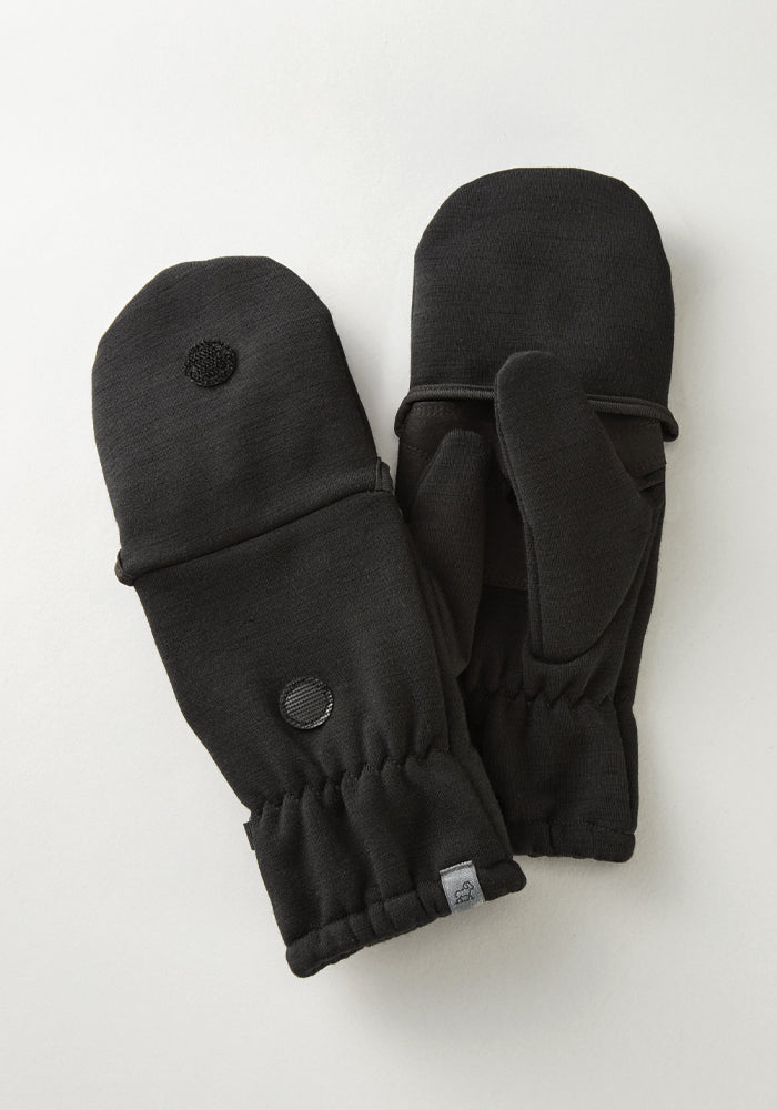 A pair of Cameron Glove Mittens from Woolx, in black, rests on a white surface. They have elastic cuffs and a small round patch on the back of each mitten, made from a soft Merino Wool Blend for enhanced warmth.