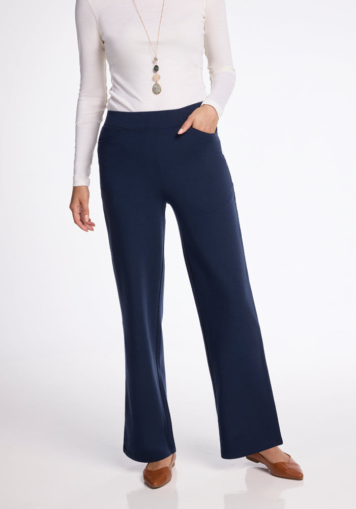 A woman is wearing a white long-sleeve top, Ellie Wide Leg Pants by Woolx in Deep Navy, and brown flat shoes. With her left hand in her pants pocket and adorned with a long necklace, she stands gracefully against a plain white background.