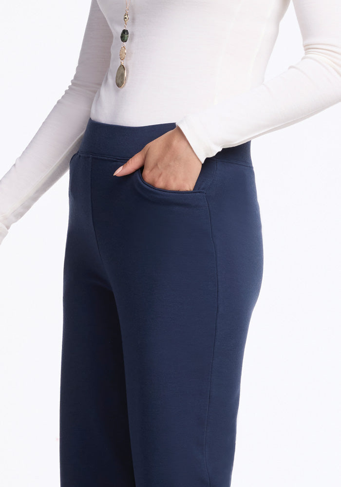 A person wearing a white long-sleeve top and Ellie Wide Leg Pants in Deep Navy from Woolx, with their hand in the pocket. The high-waisted, wide-leg silhouette enhances their look, and they are also adorned with a long necklace featuring multiple pendants.