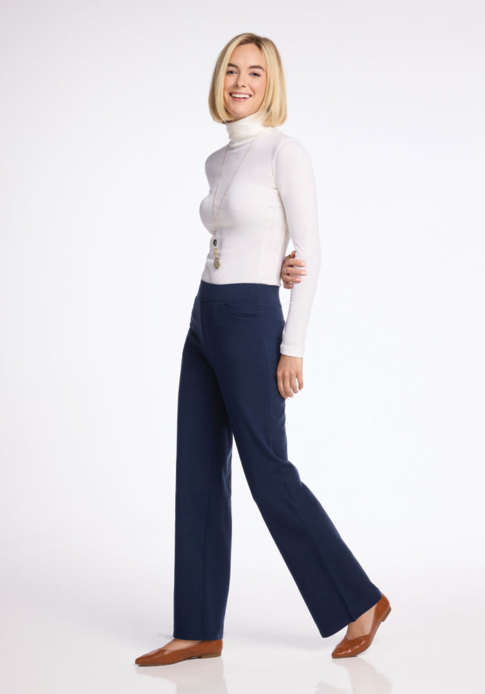 A person with a light bob hairstyle is smiling and standing, wearing a white turtleneck paired with Woolx's Ellie Wide Leg Pants in Deep Navy. They are accessorized with a long necklace and brown shoes, posed against a light background.