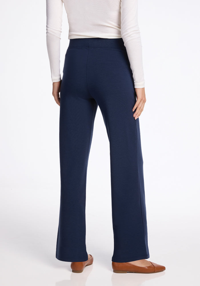Rear view of a person wearing a white long-sleeve top, high-waisted Ellie Wide Leg Pants in deep navy by Woolx, and brown flat shoes, standing against a plain white background.