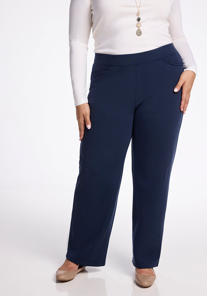 A person is wearing a long-sleeved white top paired with the Ellie Wide Leg Pants - Deep Navy by Woolx, standing against a white background. Their outfit features high-waisted pants complemented by a long necklace and beige shoes.