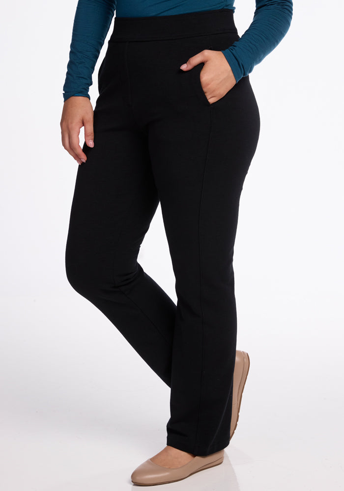 A person is wearing Woolx's Emerson Straight Leg Pants, renowned for their comfort as work pants. Paired with a long-sleeve blue top, they have one hand in their pocket and are sporting tan-colored shoes. The white background accentuates the sleek appearance of the black pants crafted from soft Merino wool. | Sarah is 5'8", wearing a size S