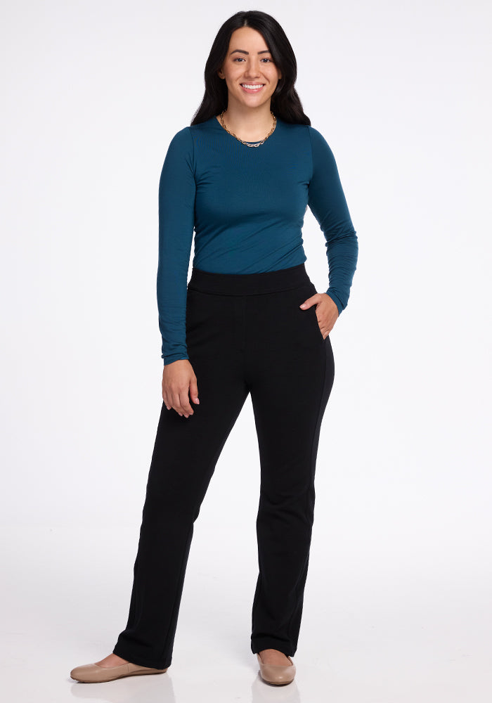 A person with long dark hair smiles while wearing a blue long-sleeve top and the sleek Emerson Straight Leg Pants in black from Woolx. They stand against a plain white background, with one hand in a pocket and the other resting at their side, highlighting their light-colored shoes made of Merino wool.