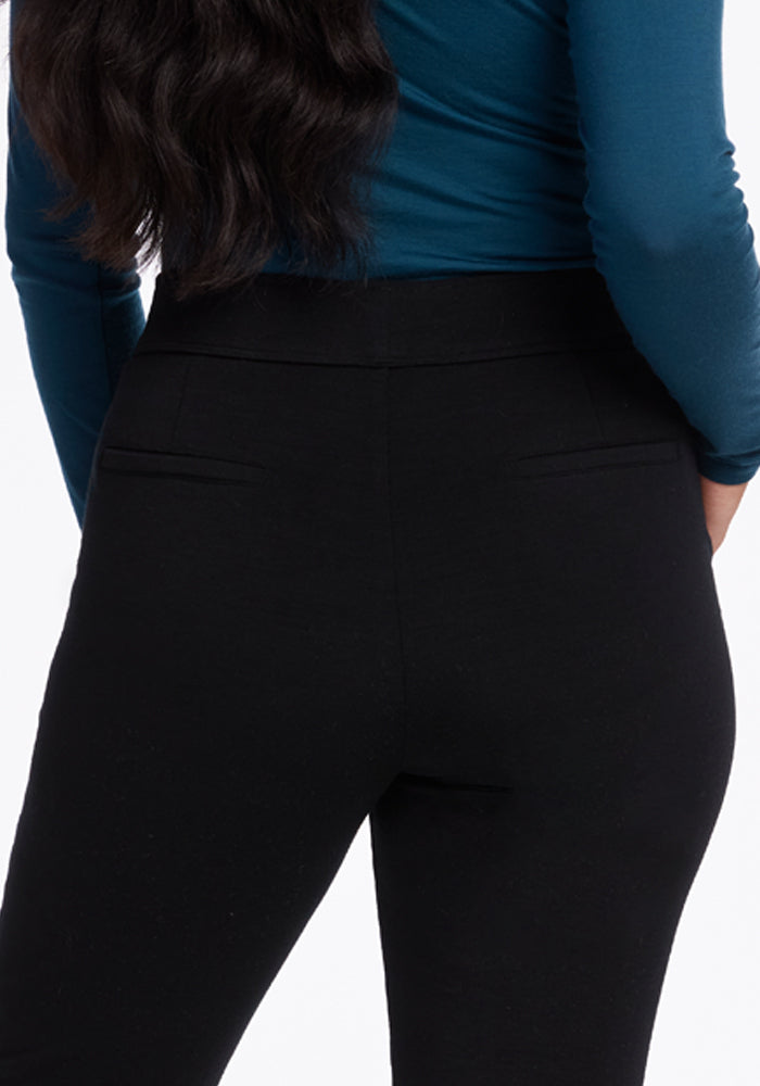 A person is standing with their back to the camera, wearing a blue long-sleeve top and sleek Woolx Emerson Straight Leg Pants in black. Their hair is long and wavy, set against a plain white background.