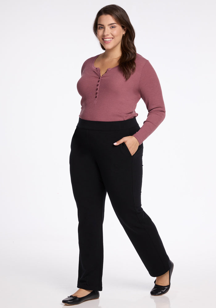 A woman in a long-sleeve, button-up maroon top and sleek Emerson Straight Leg Pants by Woolx stands with her hand in her pocket. She is smiling against a plain white background and wearing black ballet flats.