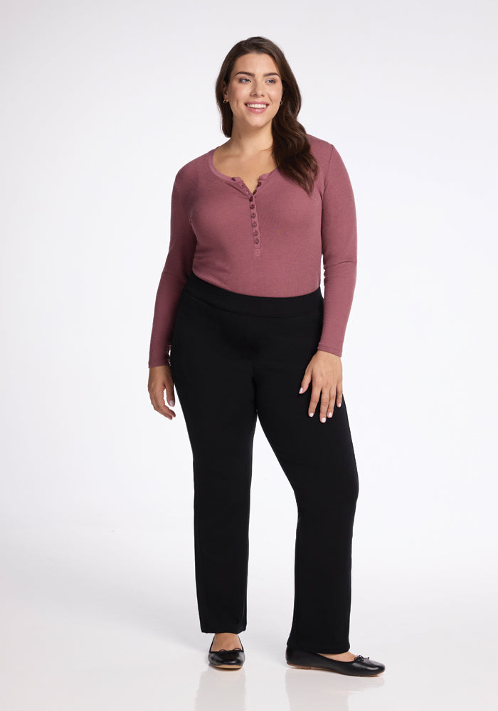 A woman with long dark hair smiles while standing. She is wearing a sleek straight-leg fit pair of Woolx's Emerson Straight Leg Pants in black, complemented by a maroon, long-sleeved top and black flats against a plain white background.