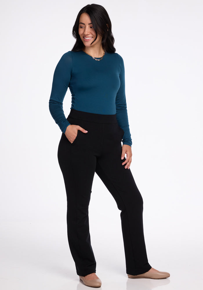 A person with long dark hair is smiling and standing against a plain background, exuding a sleek professional look. They are wearing a teal long-sleeve top paired with petite Emerson Straight Leg Pants in black from Woolx, complemented by beige shoes. With one hand casually placed in a pocket, they appear relaxed and confident.