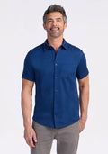 A smiling man with short dark hair and a beard wears the Woolx Endi Full Button Down Shirt in Estate Blue, paired with gray pants, standing against a plain white background. | Matthew is 6’, wearing a size L