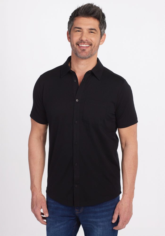 A man with short hair and a beard smiles while standing, wearing the versatile Woolx Endi Full Button Down Shirt in black made from Merino wool, paired with blue jeans against a plain white background. | Matthew is 6’, wearing a size L