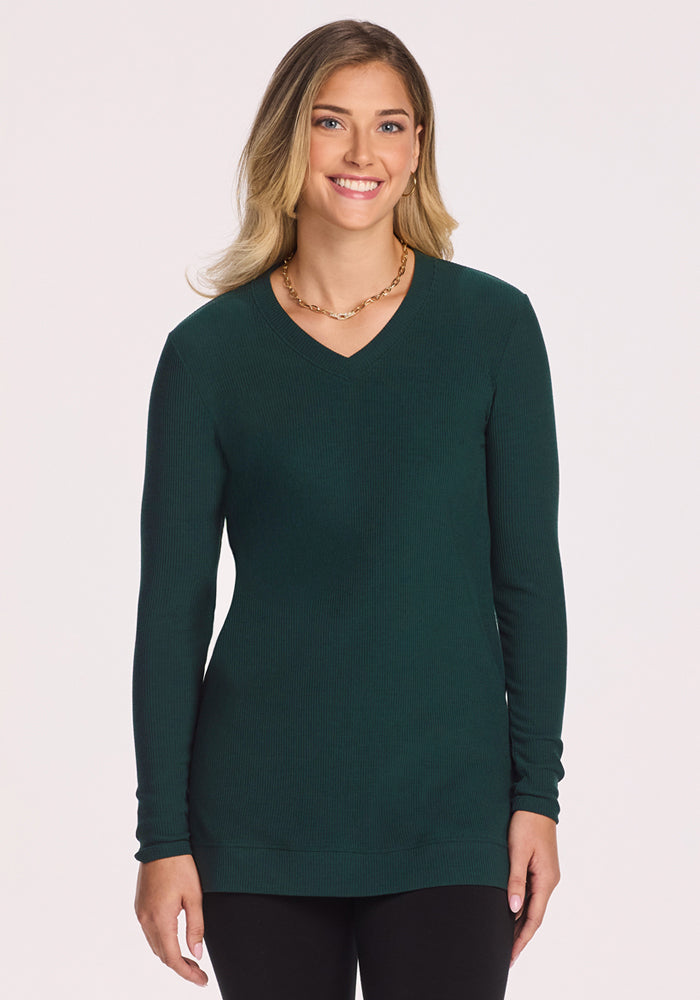 A woman with long blonde hair smiles facing forward, wearing the Woolx Evie Ribbed Tunic in Deep Timber and a gold necklace, posed against a plain light background.