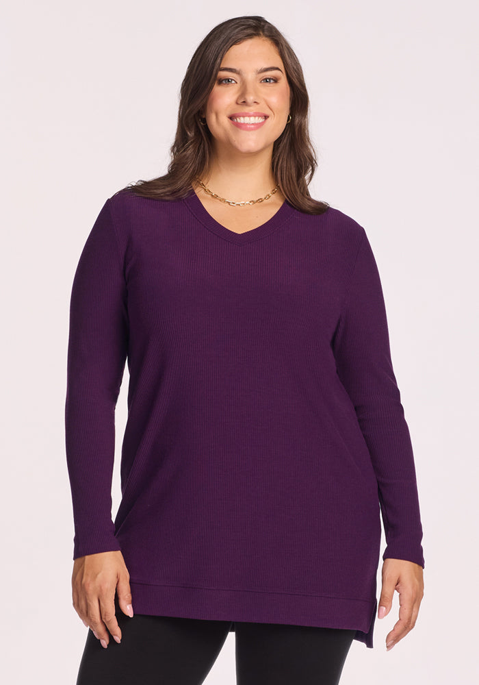 A woman with long brown hair smiles as she wears the Woolx Evie Ribbed Tunic in Majestic Purple, a cozy merino wool v-neck sweater, paired with black leggings. She stands against a plain light background.