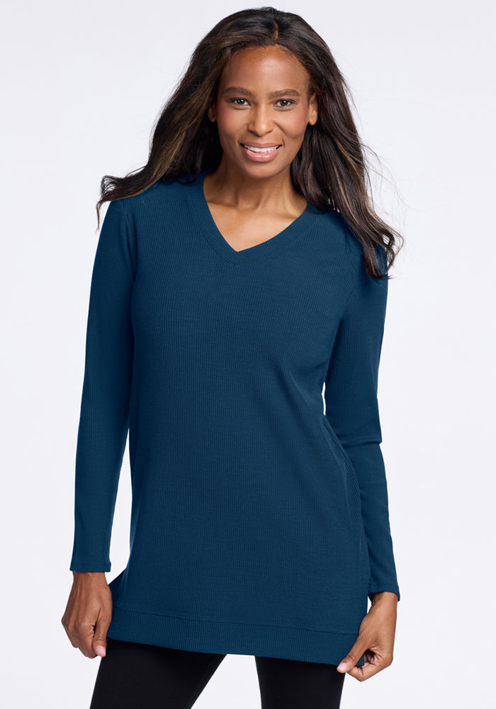 A woman with long dark hair smiles, wearing the Woolx Evie Ribbed Tunic in Majolica Blue—long-sleeved and V-neck—paired with black leggings against a plain white background. Final Sale.