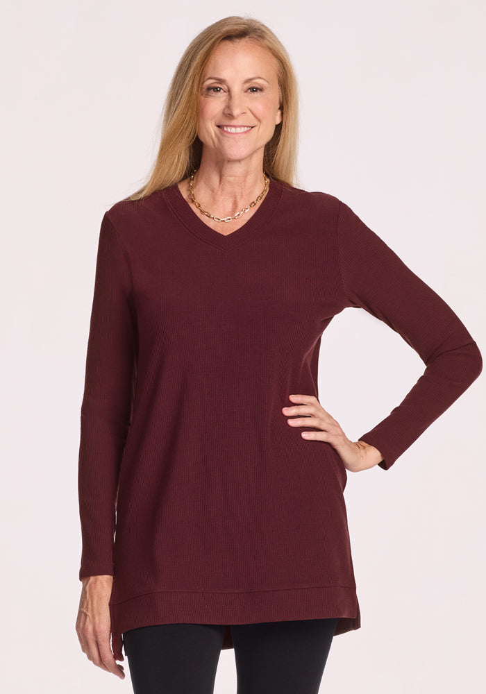 A woman with long blonde hair smiles with one hand on her hip, wearing the Woolx Evie Ribbed Tunic in Toasted Garnet (Final Sale) and black leggings, posed against a plain light background.