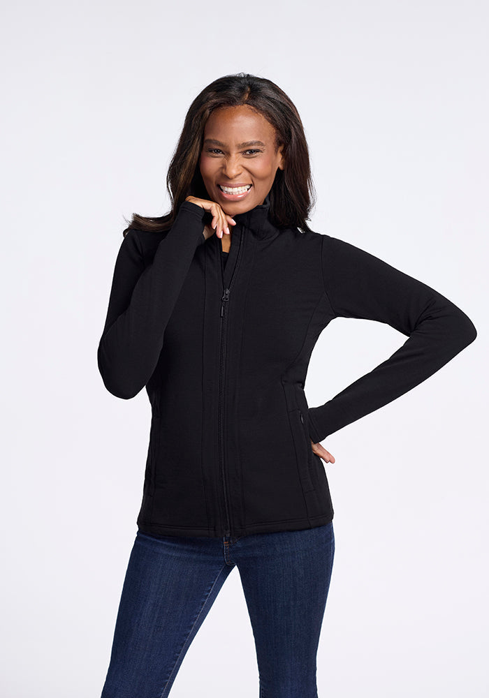 A woman with long hair is smiling and posing against a plain white background. She is wearing the Finley Zip Up Sweatshirt in black from Woolx, made of luxurious Australian merino wool, paired with blue jeans. With her left hand on her hip and her right hand touching her face, she effortlessly showcases the flattering look provided by the no-hood design of the sweatshirt. | Keesha is 5’9.5”, wearing a size S