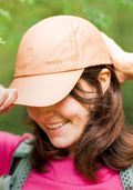 A person wearing the Woolx Frankie Baseball Hat in Salmon smiles while adjusting the brim. The adjustable merino wool cap sits over brown hair, paired with a pink top and green backpack straps against a softly blurred green background.
