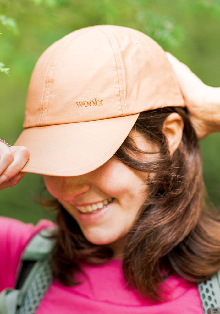 A person wearing the Woolx Frankie Baseball Hat in Salmon smiles while adjusting the brim. The adjustable merino wool cap sits over brown hair, paired with a pink top and green backpack straps against a softly blurred green background.