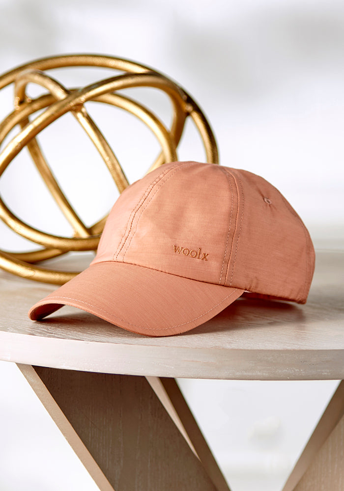 The Frankie Baseball Hat, in peach merino wool with "Woolx" embroidery, sits on a round wooden table. Its adjustable strap ensures a perfect fit. A gold geometric sculpture elegantly decorates the background.