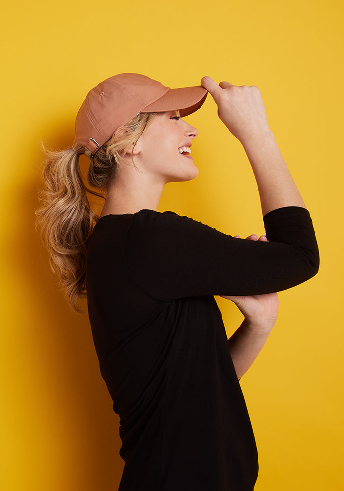 A person wearing a Woolx Frankie Baseball Hat smiles sideways against a bright yellow background. Their black shirt and ponytail complement the brown merino wool cap, and they touch its adjustable strap.