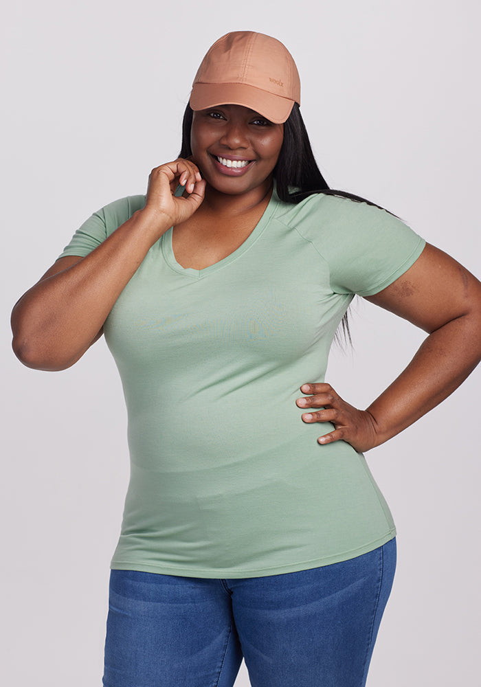 A woman with long dark hair smiles at the camera, wearing a green V-neck shirt, blue jeans, and a Woolx Frankie Baseball Hat with an adjustable strap. One hand rests on her hip while the other touches her face, posing against a plain light background.