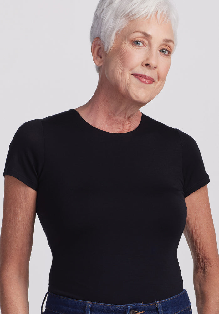 An older adult with short white hair is wearing a black Gracie Bodysuit by Woolx and dark blue jeans. They stand against a plain light background, looking at the camera with a slight smile.