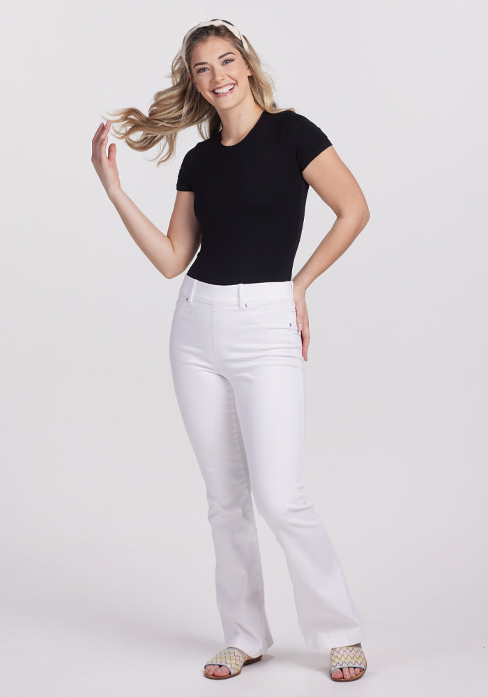 A woman in a chic Gracie Bodysuit - Black by Woolx beams, striking a pose with one hand in her hair. She enhances her outfit with white jeans, sandals, and a headband against a minimalist white backdrop.