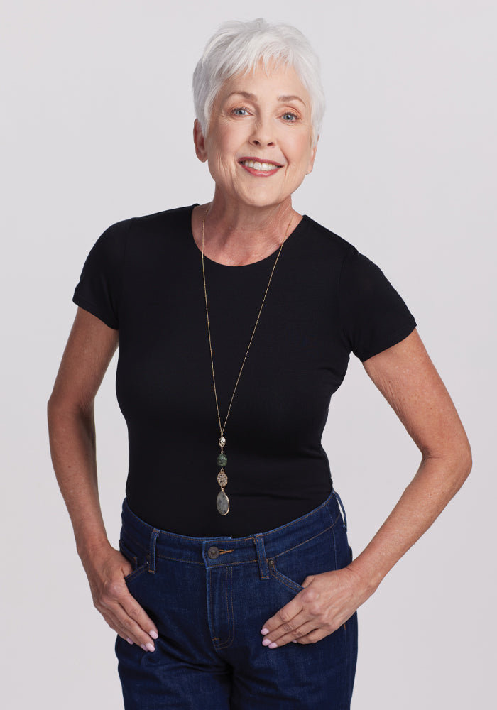 An older woman with short white hair smiles confidently, wearing the Woolx Gracie Bodysuit - Black, blue jeans, and a long necklace with a pendant. She stands with her hands in her pockets against a plain grey background. | Kathy is 5’9”, wearing a size S