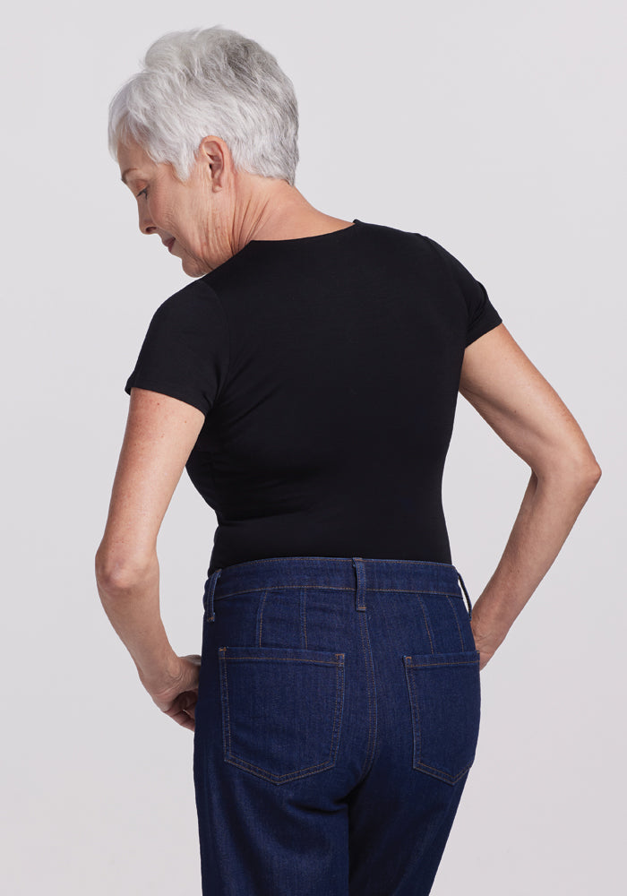 An older adult with short gray hair wears a black Gracie Bodysuit from Woolx and blue jeans, standing with their back turned. They glance over their shoulder, hands on hips against a plain backdrop, exuding casual comfort and style.