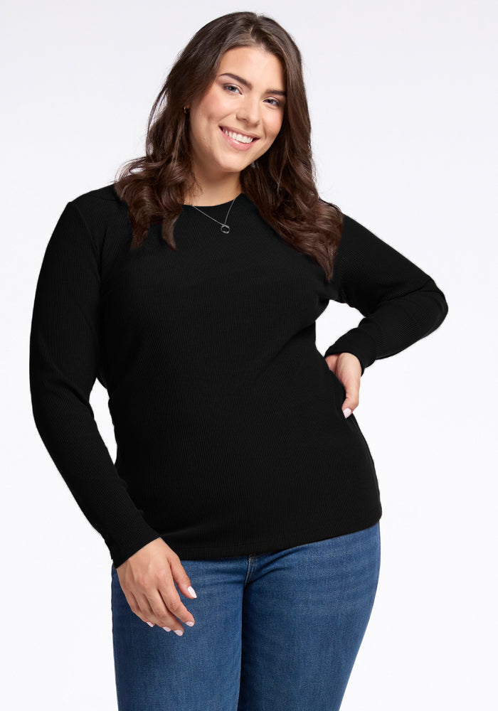 A person with long brown hair smiles at the camera, wearing the Woolx Hadley Ribbed Crew in black and blue jeans. They have one hand on their hip and the other arm relaxed by their side, exuding a luxurious feel against the plain white background. | Juliana is 5'10", wearing a size XL