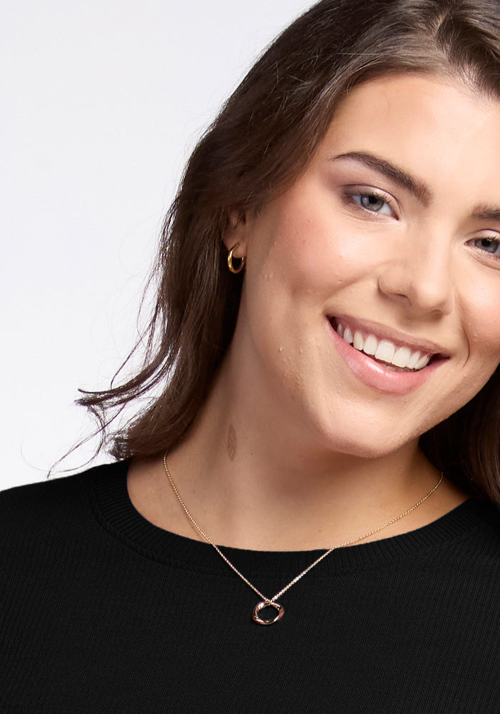 A smiling person with long hair is wearing the Hadley Ribbed Crew in black by Woolx, which exudes a luxurious feel. They pair it with gold hoop earrings and a necklace featuring a circular pendant. The image beautifully captures the elegance of their face and shoulders.